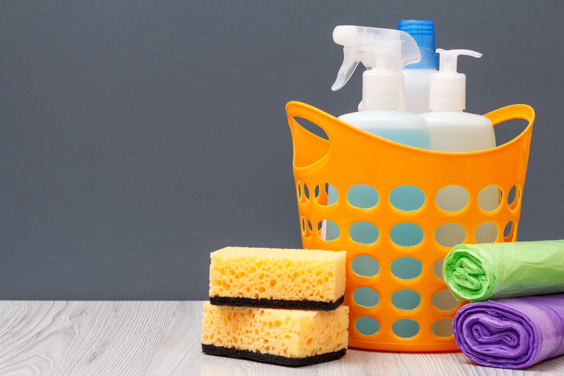 Botellas de líquido para lavar platos en una cesta y esponjas, bolsas de basura sobre fondo gris.